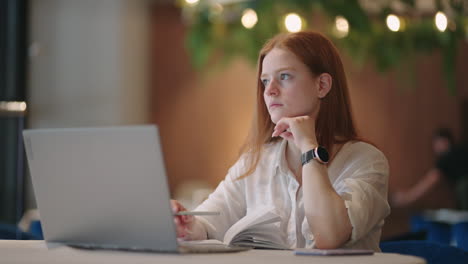 thoughtful-brooding-remote-working-red-haired-woman-sitting-infront-of-a-laptop-or-notebook-in-casual-outfit-on-her-work-desk-in-her-modern-airy-bright-living-room-home-office-with-many-windows